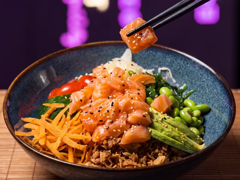 Photo d'un poke bowl saumon, edamame, avocat et wakame, un cube de saumon levé aux baguettes chez Yushithaï, le meilleur poke bowl à Lyon 6, Brotteaux, près du métro Masséna