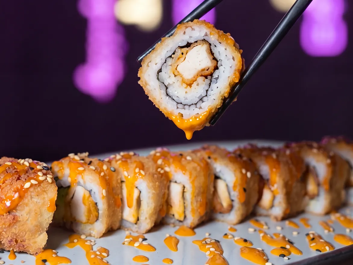Image d'un california pané au poulet et sauce spicy mayo levé entre deux baguettes, devant les néons roses de Yushithaï, le meilleur restaurant japonais halal à Lyon 6, Masséna, près du métro Brotteaux
